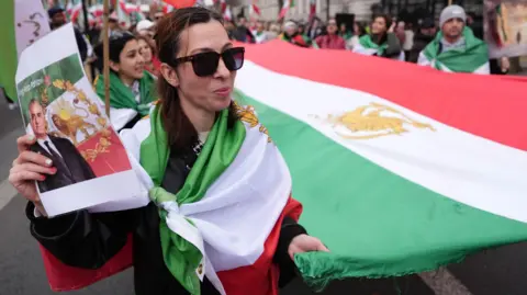 British Iranians take to streets of Manchester hours after US-Israeli strikes PA Media A woman smiles as she and others hold the sides of a large version of the Iranian flag with a Persian symbol in the centre. She also holds a printout of former Shah (king) Reza Pahlavi.