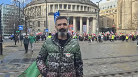 British Iranians take to streets of Manchester hours after US-Israeli strikes Dainel poses in front of the rally outside the circular Manchester Central Library. He wears a zipped up coat with an Iranina badge and flag flying from his pocket.
